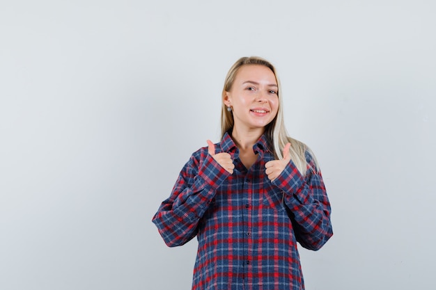 Mujer rubia en camisa a cuadros que muestra los pulgares dobles hacia arriba y luciendo atractiva, vista frontal.