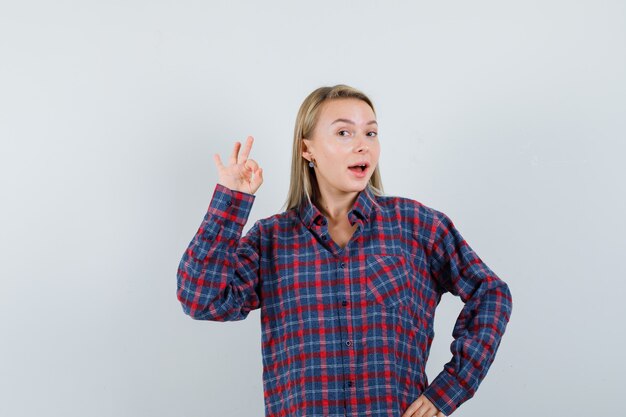 Mujer rubia en camisa a cuadros que muestra el signo de ok y parece feliz, vista frontal.