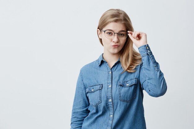 Mujer rubia en camisa de mezclilla y gafas, frunce ligeramente los labios, sueña con ropa nueva, aislado con espacio de copia para publicidad o texto promocional.