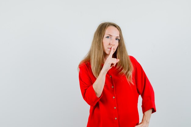 Mujer rubia en camisa roja mostrando gesto de silencio y mirando con cuidado,