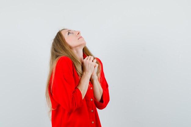 Mujer rubia en camisa roja uniendo las manos en gesto de oración y mirando esperanzado,