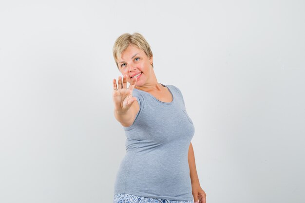 Mujer rubia en camiseta azul claro posando y levanta una mano y luciendo optimista, vista frontal.