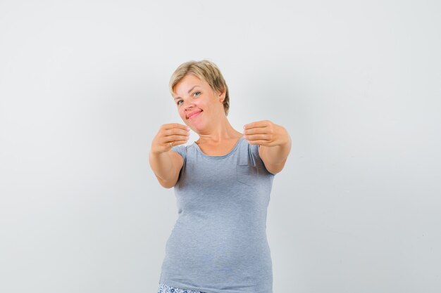 Mujer rubia en camiseta azul claro que invita a venir y que parece feliz, vista frontal.