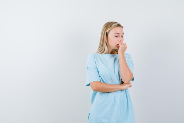 Mujer rubia en camiseta azul mordiendo el puño, sosteniendo la mano debajo del codo y mirando ansiosa