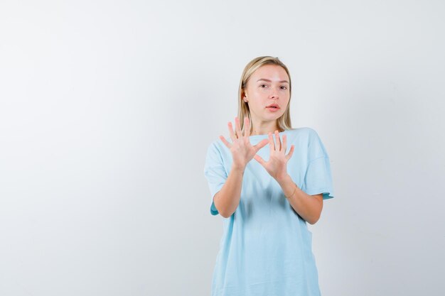 Mujer rubia en camiseta azul mostrando gesto de restricción y mirando asustada