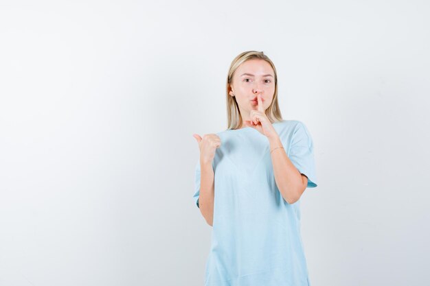 Mujer rubia en camiseta azul mostrando gesto de silencio, apuntando a un lado con el pulgar