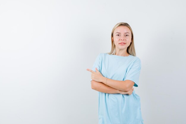 Mujer rubia en camiseta azul de pie con los brazos cruzados, apuntando hacia la izquierda con el dedo índice