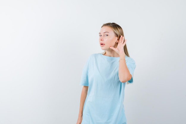 Mujer rubia en camiseta azul sosteniendo la mano cerca de la oreja para escuchar