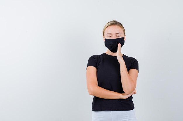 Mujer rubia en camiseta negra, pantalón blanco, máscara negra sosteniendo una mano cerca de la boca