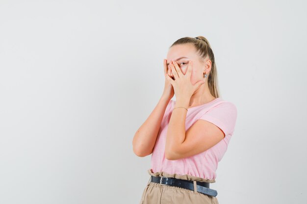 Mujer rubia en camiseta, pantalones mirando a través de los dedos y luciendo delicada, vista frontal.