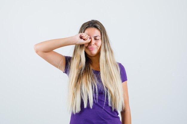 mujer rubia en camiseta violeta frotándose los ojos mientras llora y parece ofendida, vista frontal.