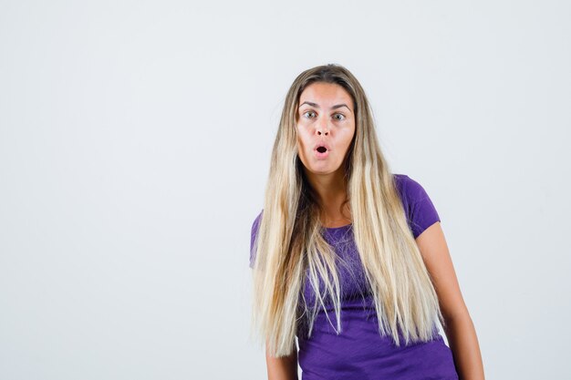 mujer rubia en camiseta violeta mirando al frente y mirando sorprendida, vista frontal.