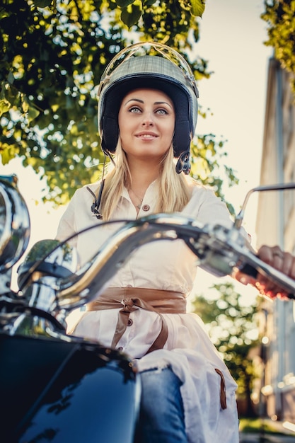 Mujer rubia con casco de moto sentada en moto scooter sobre hojas verdes y sol.