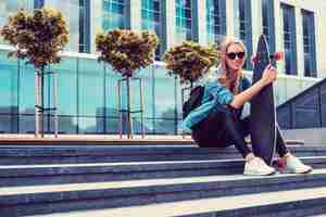 Foto gratuita mujer rubia casual con camisa vaquera posando en las escaleras con longboard sobre un edificio de cristal.