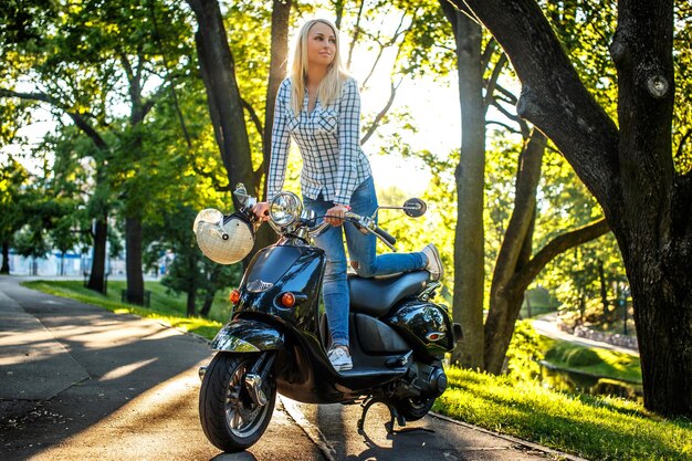 Foto gratuita mujer rubia casual en jeans azules y camiseta blanca posando en el parque cerca de scooter azul.