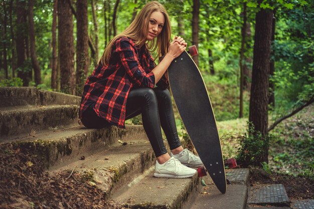 Mujer rubia casual posando con longboard en las escaleras en un parque natural salvaje.