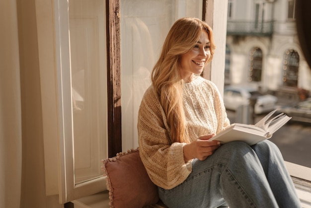 Mujer rubia caucásica adulta con suéter cálido lee un libro mientras se sienta en el alféizar de la ventana en la habitación Concepto de descanso y recuperación
