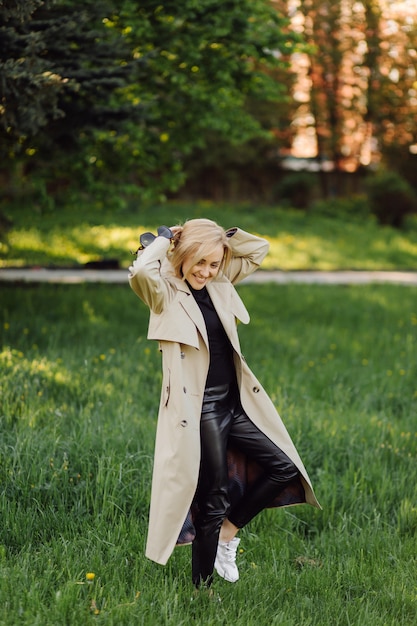 Mujer rubia caucásica wearind trinchera sonríe felizmente en un día soleado de primavera afuera caminando en el parque