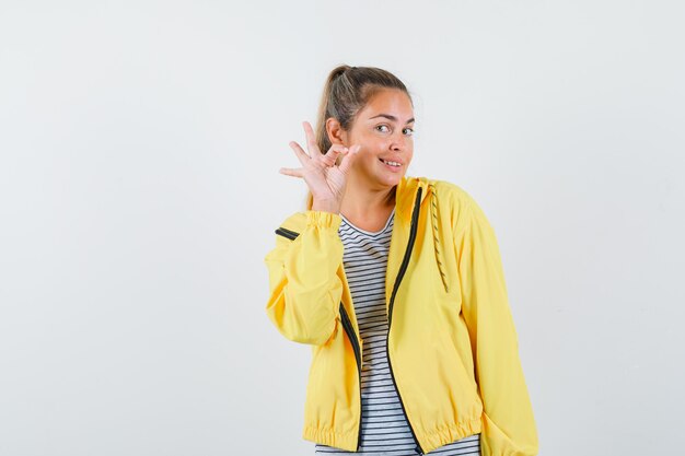 Mujer rubia en chaqueta de bombardero amarilla y camisa a rayas que muestra el signo de ok y se ve bonita