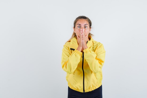 Mujer rubia en chaqueta de bombardero amarilla y pantalón negro de pie en gesto de oración y mirando sorprendido