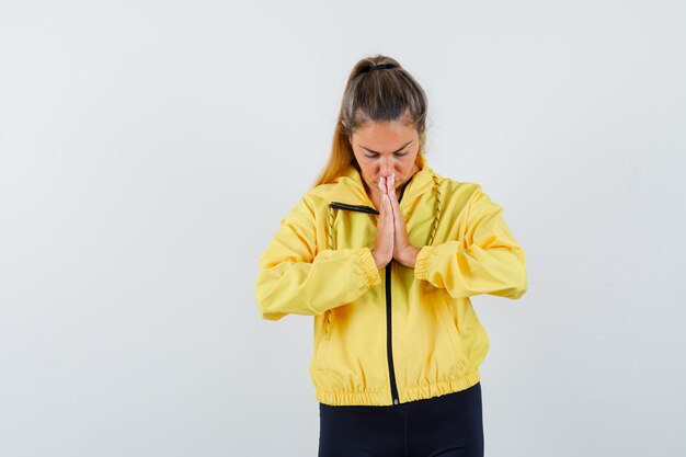 Mujer rubia en chaqueta de bombardero amarilla y pantalón negro de pie en pose de oración y mirando enfocado