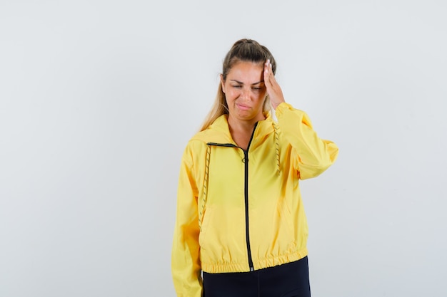 Mujer rubia en chaqueta de bombardero amarillo y pantalón negro con dolor de cabeza y mirando molesto