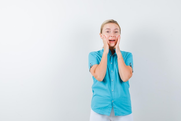 Mujer rubia cogidos de la mano en las mejillas en blusa azul y mirando feliz aislado