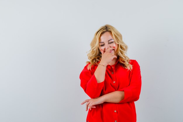 Mujer rubia cubriendo la boca con las manos, sonriendo en blusa roja y mirando feliz