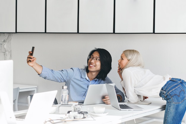 Mujer rubia delgada con camisa blanca y jeans posando junto a la mesa mientras su colega asiático haciendo selfie. Inoor retrato de trabajador chino en vasos divirtiéndose con la secretaria.