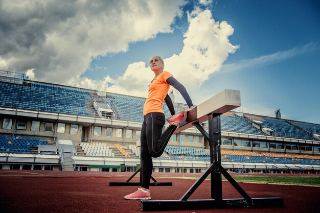 Mujer rubia delgada en ropa deportiva haciendo ejercicios en el estadio bajo un cielo azul con nubes blancas.