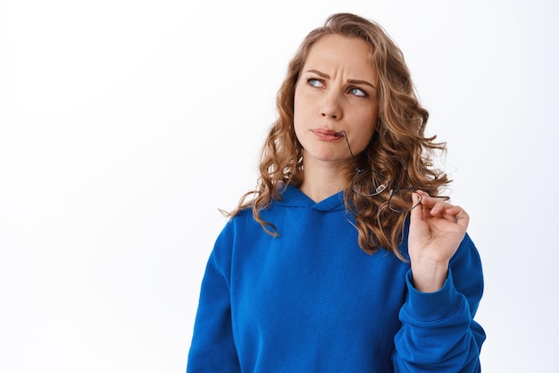 Mujer rubia desconcertada mordiendo anteojos y frunciendo el ceño pensativa mirando a un lado con cara pensativa haciendo elección teniendo dudas de pie sobre fondo blanco