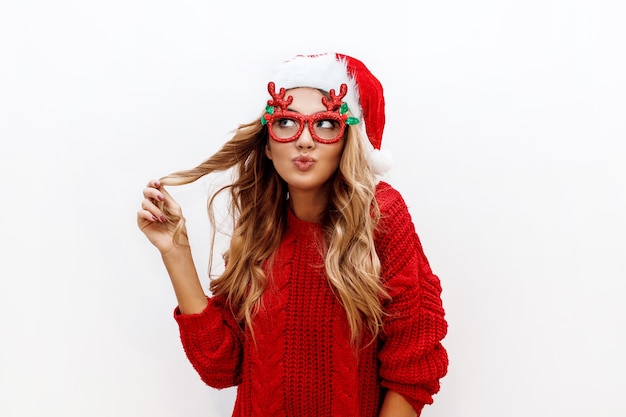 Mujer rubia despreocupada alegre en gafas de mascarada lindo y sombrero de año nuevo en suéter de punto rojo posando en la pared blanca. Aislar. Navidad y nuevo concepto de fiesta de prueba.