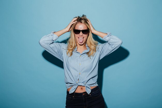 Mujer rubia divertida en camisa y gafas de sol mostrando su lengua