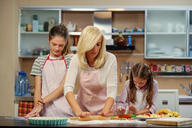 Mujer rubia y dos adolescentes que muestran cómo amasar en la cocina de la casa.