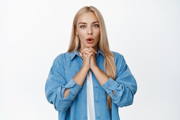 Mujer rubia emocionada jadeando por la anticipación de estar al límite con la historia mirando con interés y sorpresa de pie sobre fondo blanco.