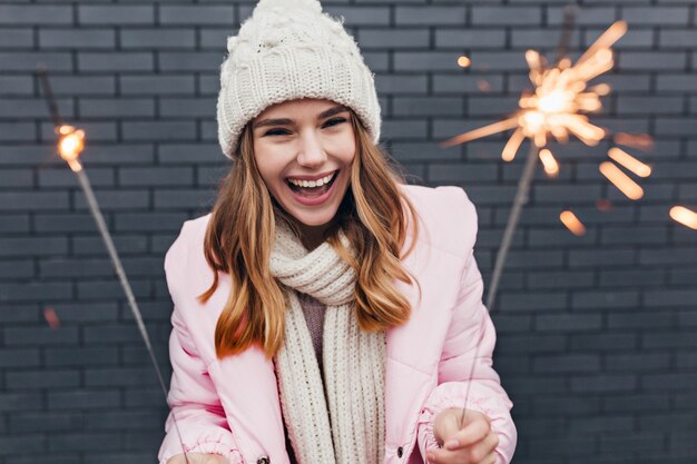 Mujer rubia emocionada en traje romántico celebrando el año nuevo. Foto al aire libre de niña alegre en gorro de punto escalofriante en Navidad.