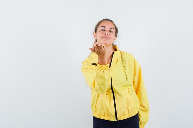 Mujer rubia enviando besos al frente con chaqueta de bombardero amarilla y pantalón negro y mirando seria