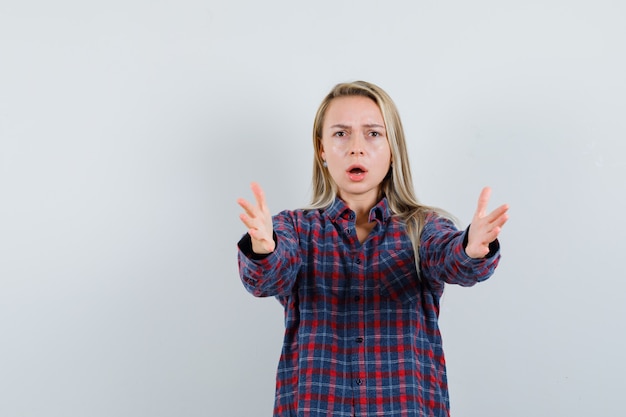 Mujer rubia estirando las manos hacia la cámara como recibiendo algo en camisa a cuadros y mirando sorprendido, vista frontal.