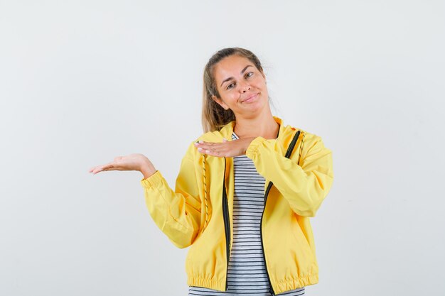 Mujer rubia estirando las manos como sosteniendo algo imaginario en chaqueta de bombardero amarilla y camisa a rayas y luciendo bonita