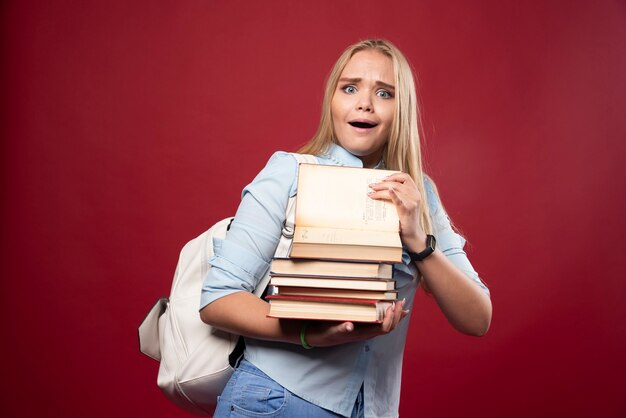 Mujer rubia estudiante sosteniendo una pesada pila de libros y parece cansada.