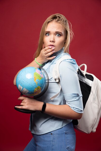 La mujer rubia estudiante sostiene un globo terráqueo y parece sorprendida.