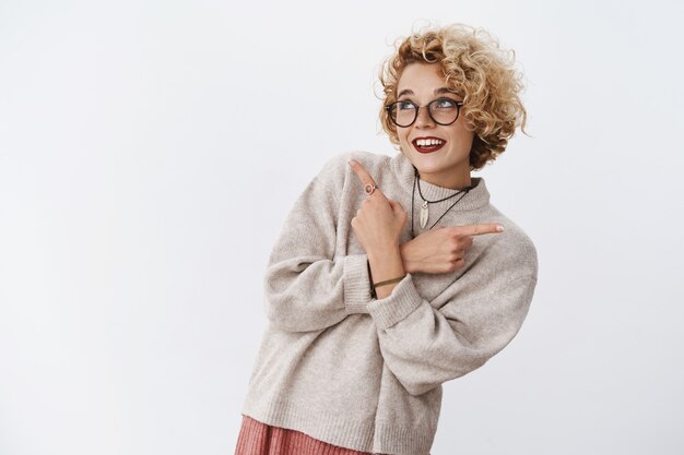 Mujer rubia europea linda soñadora y tonta que apunta hacia los lados a la izquierda y a la derecha mientras elige entre una variedad de productos geniales sonriendo ampliamente mirando hacia arriba, tomando una decisión feliz sobre la pared blanca.