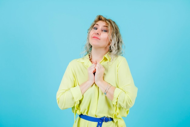 Mujer rubia feliz está mirando a la cámara sosteniendo los puños en el pecho sobre fondo azul.