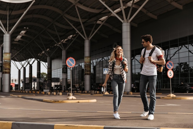 Mujer rubia feliz habla con su novio Retrato completo de una chica genial con auriculares rojos y un chico moreno con camiseta blanca camina con mochilas cerca del aeropuerto