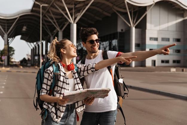 Mujer rubia feliz y hombre guapo en camisetas blancas apuntan al mismo lado Viajeros alegres con mochilas sostienen mapa cerca del aeropuerto