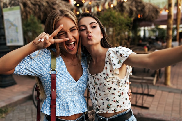 Foto gratuita mujer rubia feliz loca en blusa floral azul guiña un ojo, sonríe ampliamente y muestra el signo de la paz