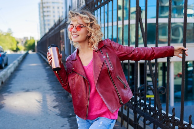 Mujer rubia feliz posando en las calles modernas, tomando café o capuchino. Elegante traje de otoño, chaqueta de cuero y suéter de punto. Gafas de sol rosas