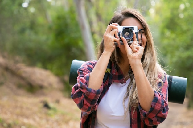 Mujer rubia feliz tomando fotos de la naturaleza con cámara y sonriendo. Viajero caucásico de pelo largo caminando o caminando en el bosque. Concepto de turismo, aventura y vacaciones de verano.