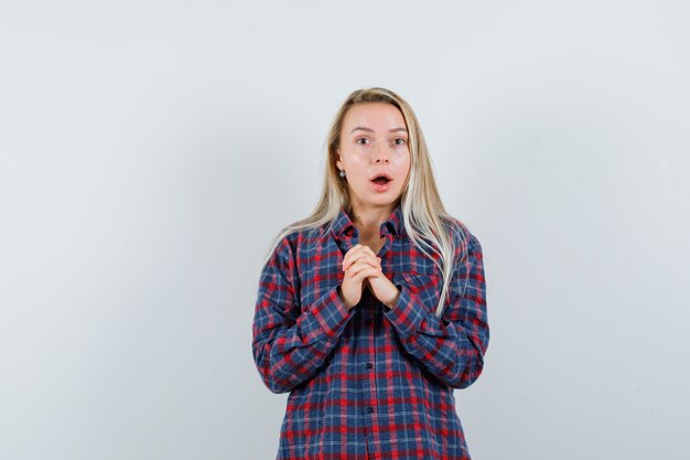 Mujer rubia frotándose las manos, de pie con la boca abierta en camisa a cuadros y mirando sorprendido. vista frontal.