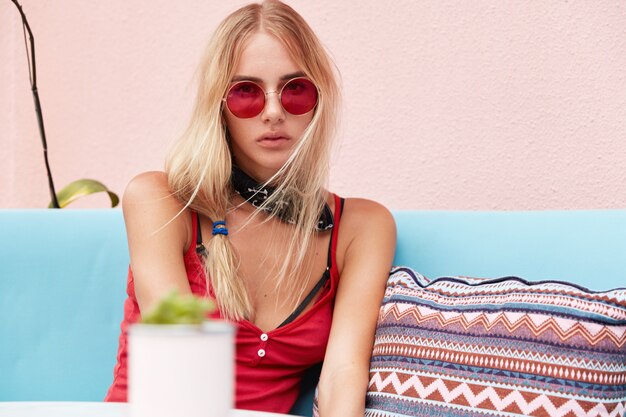 mujer rubia con gafas de sol de moda, viste ropa de moda y gafas de sol rojas, se sienta contra la pared rosa en un cómodo sofá.